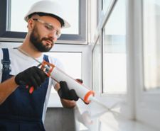 A tradesman applies sealant to a white window joint with a caulking gun. He wears gloves, goggles, and a hard hat.