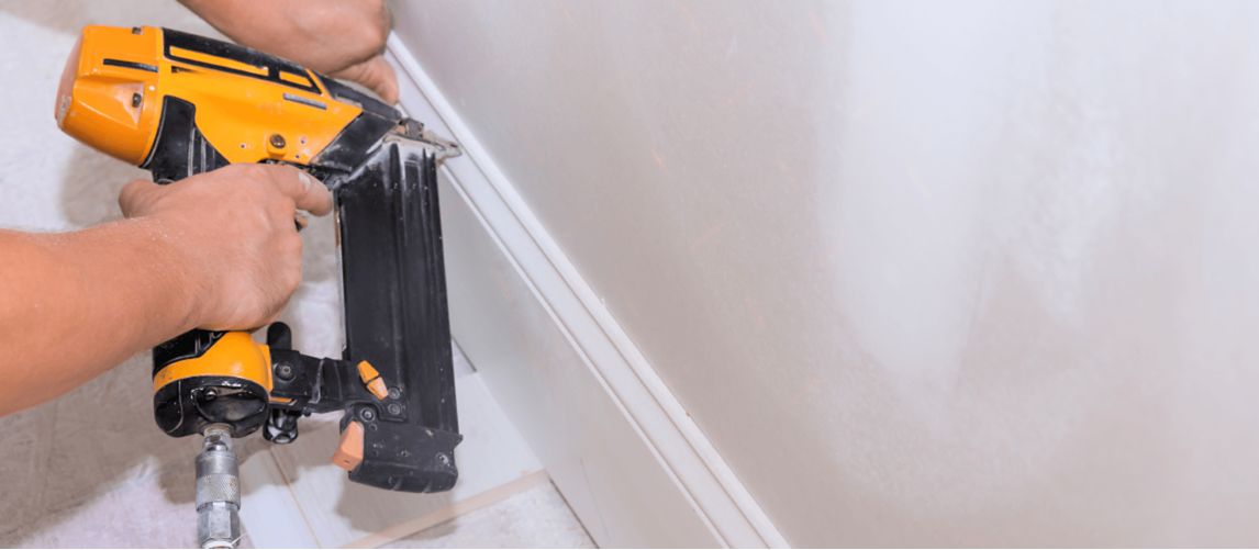 person using a nail gun to attach baseboard along a white wall
