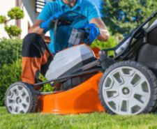 A man in a blue shirt and work gloves kneels on a green lawn, inserting a battery pack into the compartment of an orange and grey cordless lawn mower with a grass catcher attached, demonstrating a feature of a battery-powered mower for a guide to the best models.