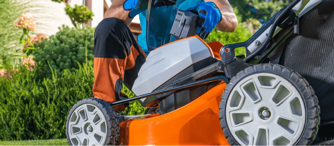 A man in a blue shirt and work gloves kneels on a green lawn, inserting a battery pack into the compartment of an orange and grey cordless lawn mower with a grass catcher attached, demonstrating a feature of a battery-powered mower for a guide to the best models.