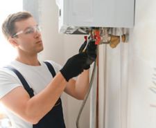 A Gas Safe engineer works on the underside of a boiler. He's wearing rubber gloves and goggles.