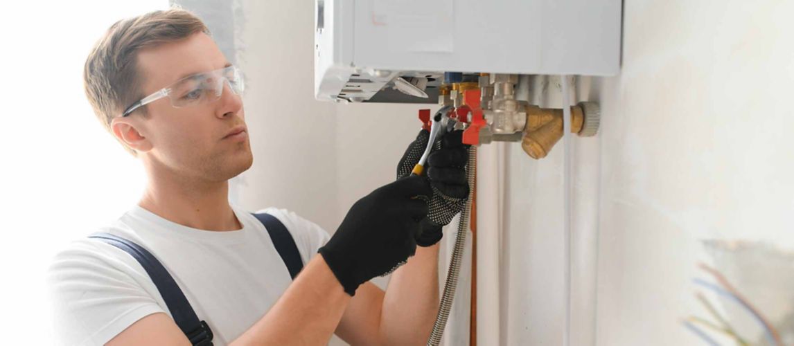 A Gas Safe engineer works on the underside of a boiler. He's wearing rubber gloves and goggles.