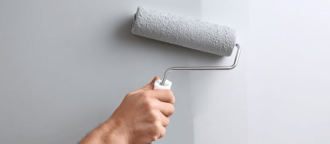Hand holding a paint roller applying light grey paint over lining paper on an interior wall