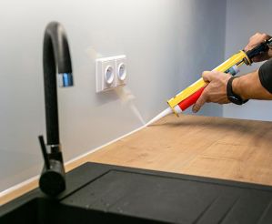 A man using a silicone caulking gun to fill gap between glass backsplash and countertop in new modern kitchen