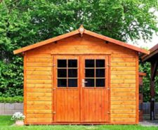 A new brown shed with an apex roof and double doors built on a grass lawn with trees in the background