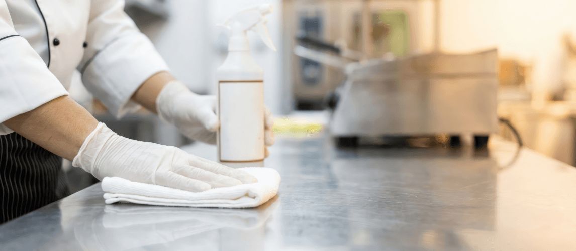 Chef wearing gloves cleaning a stainless steel kitchen counter with a spray and cloth