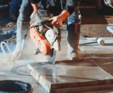 Construction worker cutting concrete paving tiles with petrol concrete cutter saw and water dust suppressor on construction site