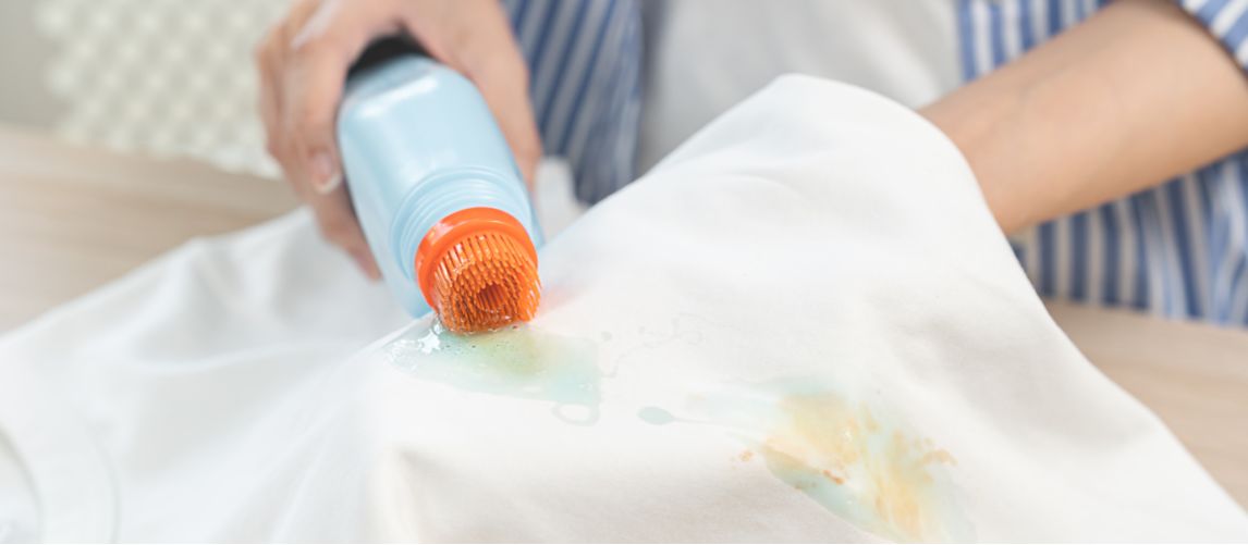 Close-up of a person removing a bleach stain from white clothes by scrubbing the yellow discoloured spot with a liquid stain remover brush.