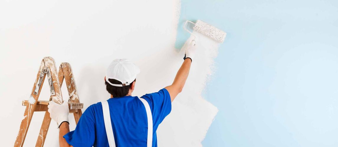 Man in overalls using a paint roller to cover a blue wall with white paint.