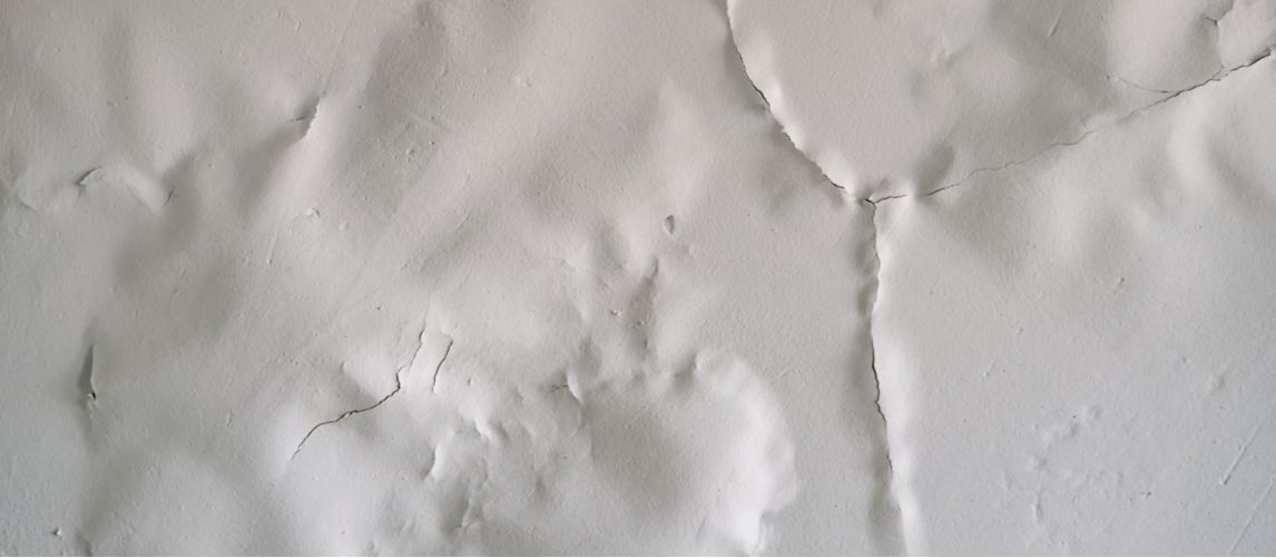 Close-up photo of white paint bubbling, blistering, and cracking on a wall surface, illustrating a common home maintenance issue caused by moisture or poor adhesion for an article on paint failure.
