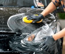 Gloved hand wiping suds off black car bonnet with yellow sponge