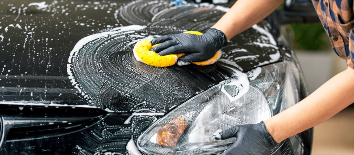 Gloved hand wiping suds off black car bonnet with yellow sponge